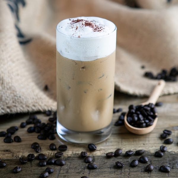 Iced coffee with whipped cream and a sprinkle of cocoa powder, surrounded by coffee beans.