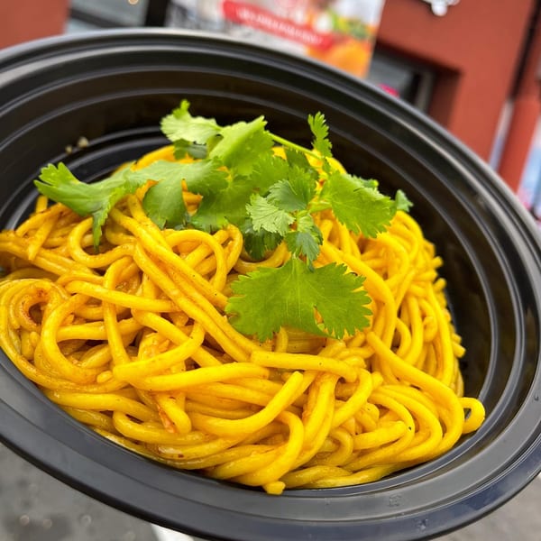 Hiccups' yellow noodles with cilantro in a black bowl, with a red building in the background.