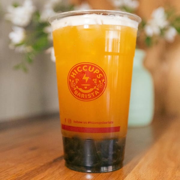 A Hiccups Barista branded plastic cup filled with a vibrant orange drink and dark boba pearls.