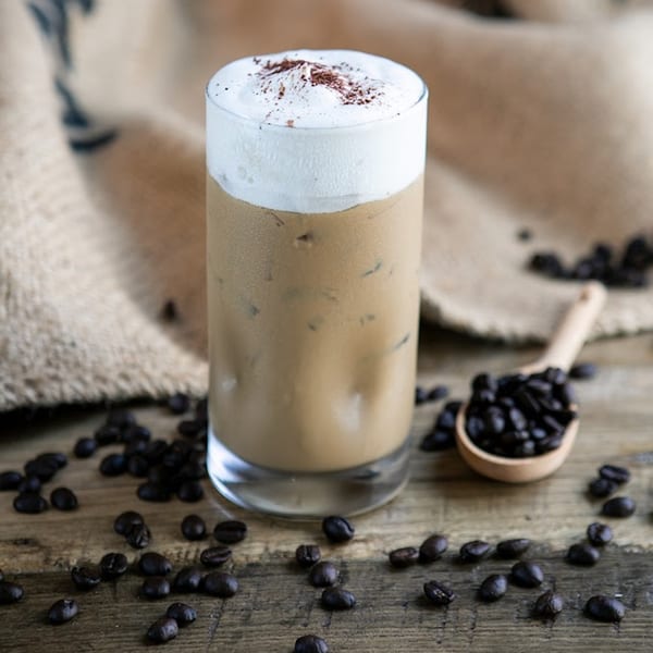 Iced coffee with whipped cream and cocoa powder, surrounded by coffee beans on a wooden surface.