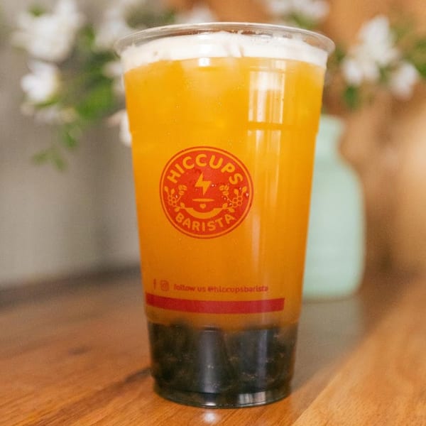 A Hiccups Barista drink, orange with dark boba, on a wooden table with blurred flowers.
