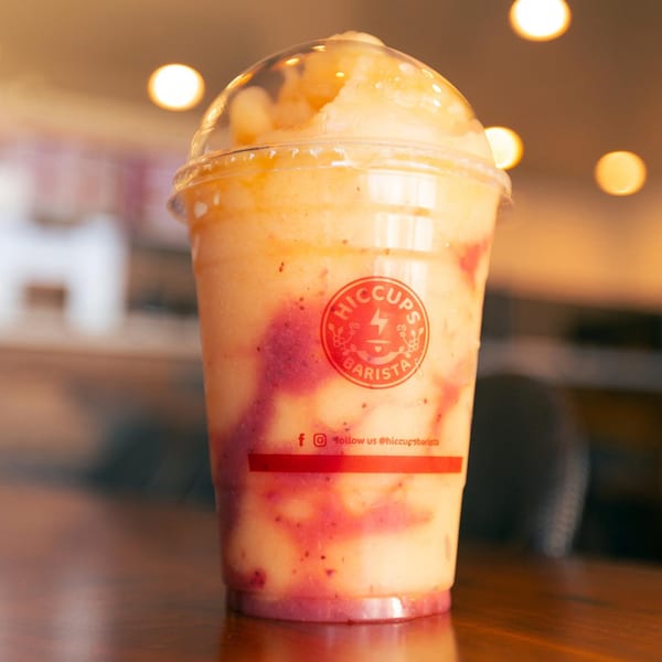 A Hiccups Barista mango and strawberry blended drink with a clear dome lid on a wooden table.