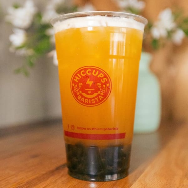 A Hiccups Barista drink, orange with dark boba, on a wooden table with blurred background.
