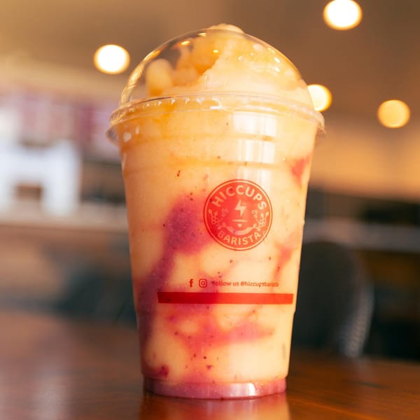 A Hiccups Barista fruit smoothie, with swirls of red and yellow, sits on a wooden table.